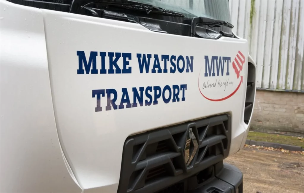 Close-up of a white lorry cab displaying the logo and branding for Mike Watson Transport (MWT), with the slogan ‘Delivered the right way’ next to a red arrow graphic
