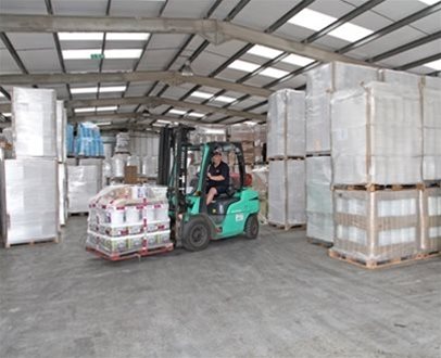 Warehouse worker operating a green forklift to move a wrapped pallet of goods between tall stacks of boxed inventory inside a spacious, well-lit storage facility.