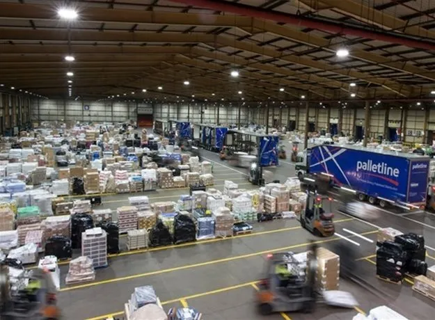 Busy Palletline distribution hub filled with stacked pallets, forklifts in motion, and branded trailers being loaded and unloaded under bright warehouse lighting.